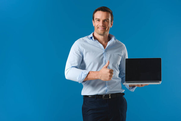 handsome man holding laptop with blank screen, showing thumb up and smiling at camera isolated on blue