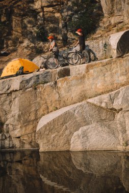 distant view of two male travelers  standing with mountain bikes near tent on rocky cliff over river clipart