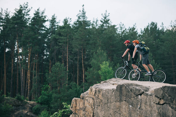 distant view of male extreme cyclists in protective helmets riding on mountain bicycles on rocky cliff in forest