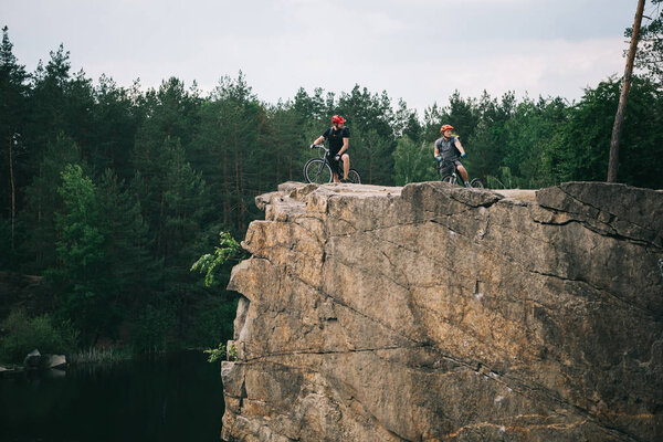 два экстремальных велосипедиста в защитных шлемах стоят с горными велосипедами на скалистой скале в лесу
