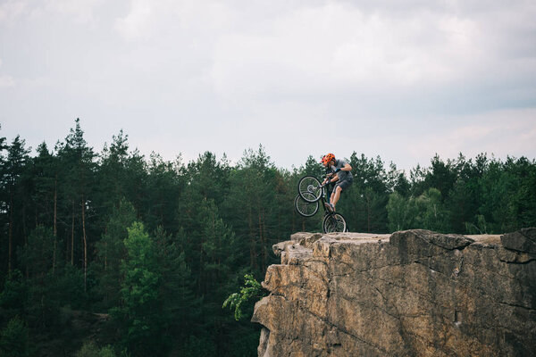 distant view of male extreme cyclists in protective helmets doing stunts on mountain bicycles on rocky cliff in forest