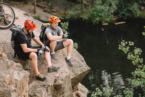 high angle view of friends cyclists with backpacks resting with sport bottle of water on rocky cliff near river in forest
