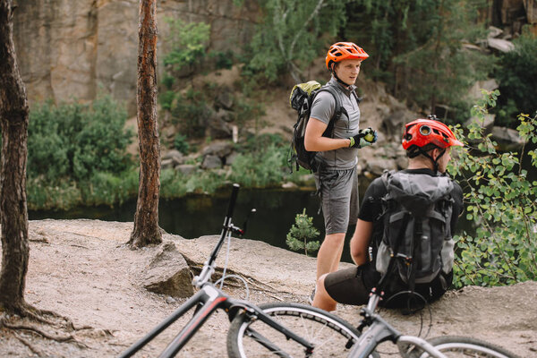 two male friends with backpacks and bicycles resting near river in forest