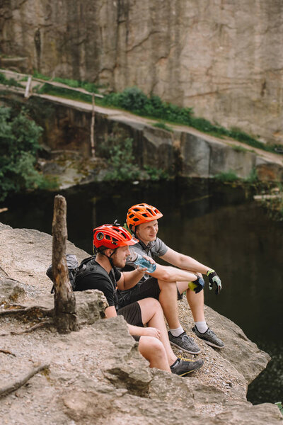 Sportsmen in protective helmets resting with apple and sport bottle of water on rocky cliff