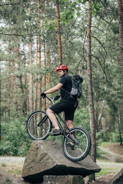 male extreme cyclist in protective helmet standing on mountain bicycle on stone in forest