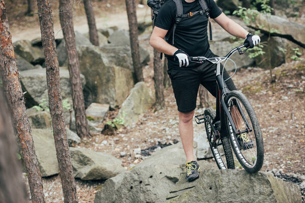 cropped image of male extreme cyclist in protective helmet standing with mountain bicycle on stone in forest