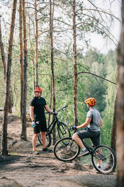 two male extreme cyclists in helmets with mountain bikes resting with sport bottles of water in forest 