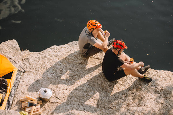 travelers in protective helmets eating canned food near tent, logs, axe and cauldron on rocky cliff over river 
