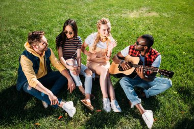 Interracial grup arkadaşıyla parkta yeşil çimenlerin üzerinde yaz günü dinlenme akustik gitar