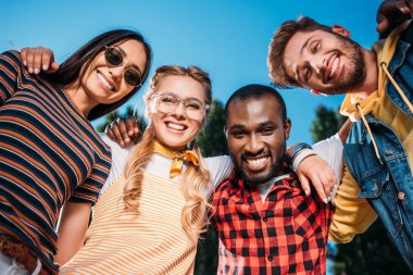 bottom view of smiling multiracial young friends looking at camera with blue sky on background clipart