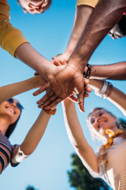 bottom view of multiracial young friends holding hands together with blue sky on backdrop clipart