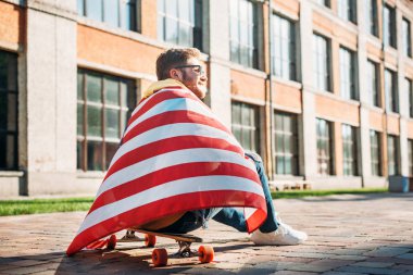 back view of young man with american flag sitting on longboard on street clipart