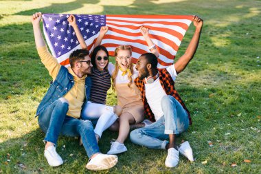 multiracial cheerful friends with american flag sitting on green grass in park clipart