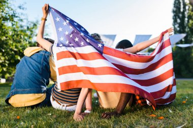 arkadan görünüşü çok ırklı arkadaşlarım parkta yeşil çimenlerin üzerinde oturan Amerikan bayrağı ile