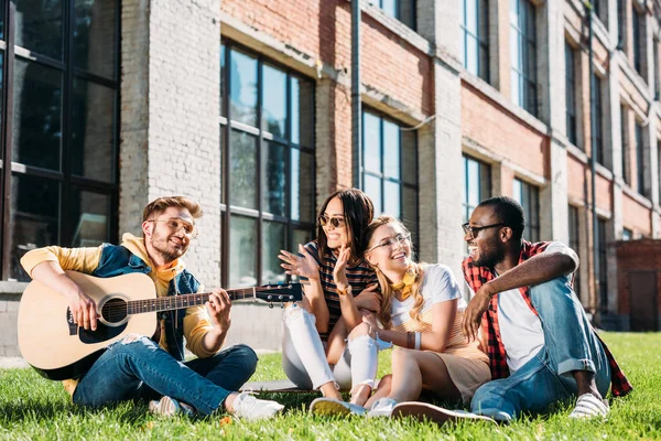 interracial group of happy friends with acoustic guitar resting on green grass on summer day