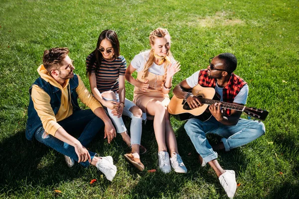interracial group of friends with acoustic guitar resting on green grass in park on summer day