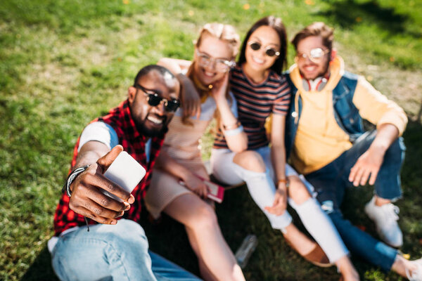 selective focus of multiethnic friends taking selfie on smartphone while resting on green lawn in park