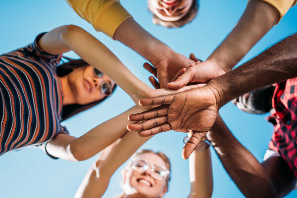 bottom view of multiracial young friends holding hands together with blue sky on backdrop