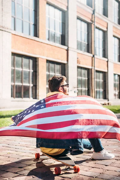 back view of young man with american flag sitting on longboard on street