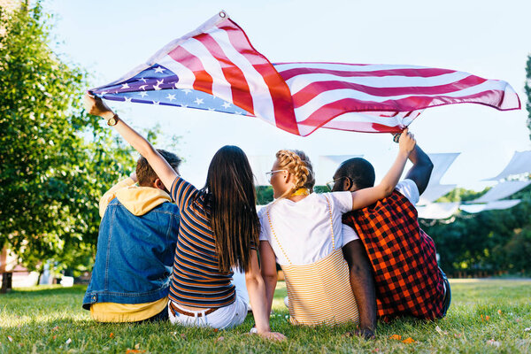 Вид сзади друзей-представителей разных рас с американским флагом, сидящих на зеленой траве в парке
