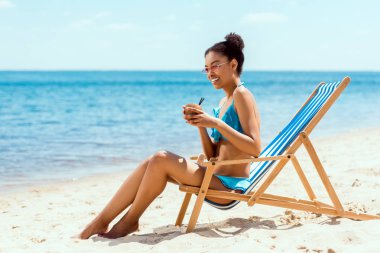 mutlu Afrikalı-Amerikalı kadın bikini ve kumsalda şezlong üzerinde otururken Hindistan cevizi kabuğu'nda kokteyl içme güneş gözlüğü