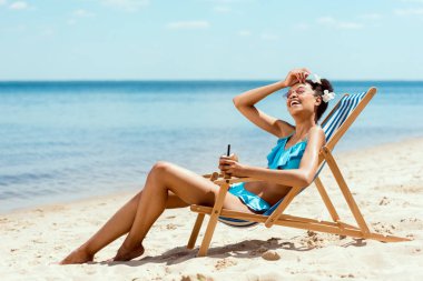 genç Afro-Amerikan kadın alın silme ve kumlu plajda şezlong üzerinde döşeme ise Hindistan cevizi kabuğu'nda kokteyl tutan bikini 