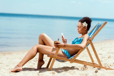 Hindistan cevizi kabuğu'nda kokteyl içme ve kumlu plajda şezlong üzerinde döşeme smartphone kullanarak kafamda çiçekli Afrikalı-Amerikalı kadın 