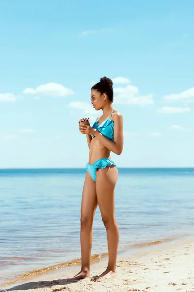 身着比基尼和太阳镜的年轻非洲裔美国妇女在沙滩上举行的椰子贝壳鸡尾酒 — 免费的图库照片