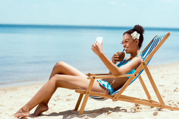 attractive african american woman with flower in head drinking cocktail in coconut shell and taking selfie on smartphone while laying on deck chair on sandy beach 