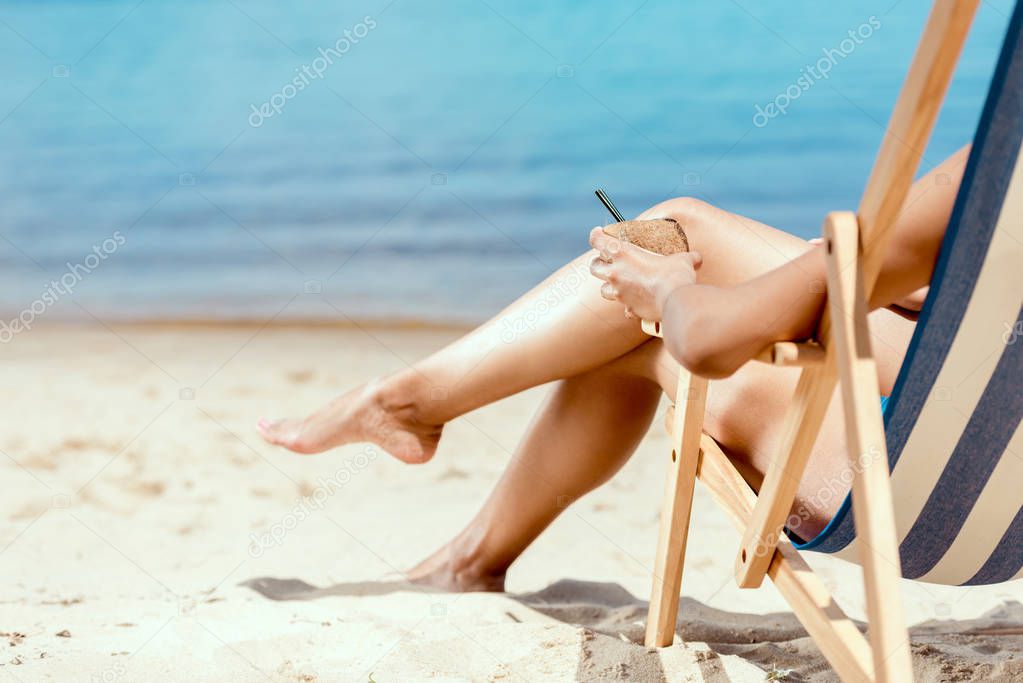 cropped image of woman holding cocktail in coconut shell and laying on deck chair on sandy beach 