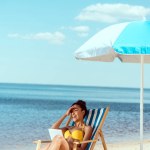 Laughing african american woman relaxing on deck chair and using digital tablet under beach umbrella in front of sea