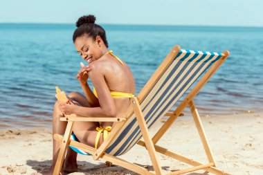 smiling african american woman applying sunscreen lotion on skin while sitting on deck chair on sandy beach  clipart