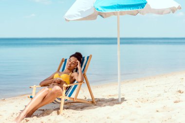 genç Afro-Amerikan kadın rahatlatıcı şezlong ve deniz önünde plaj şemsiyesi altında smartphone ile kulaklık müzik dinleme 