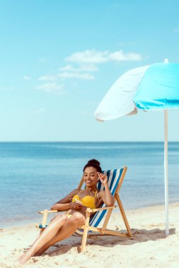 gülümseyen şezlong üzerinde rahatlatıcı ve deniz önünde plaj şemsiyesi altında smartphone ile kulaklık müzik dinleme Afrikalı-Amerikalı kadın 