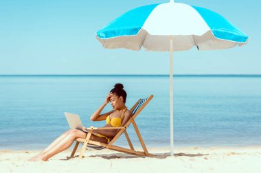 Güverte sandalyede oturan ve plaj şemsiyesi altında laptop denize cepheli kullanarak gülümseyen Afrikalı-Amerikalı kadın freelancer yan görünüm 