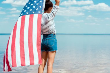 rear view of young woman holding american flag in front of sea, independence day concept clipart
