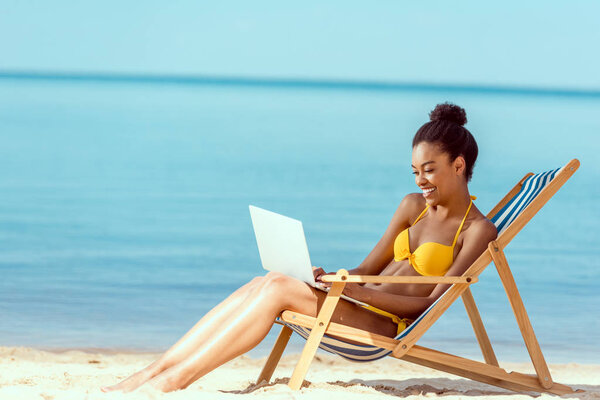 smiling african american female freelancer sitting on deck chair and using laptop on sandy beach 