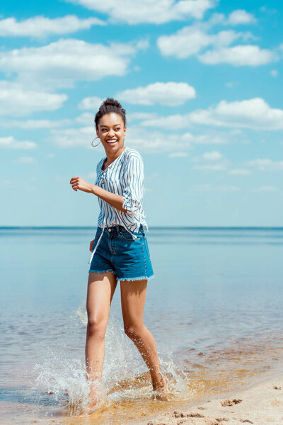 happy african american female tourist walking in sea water near sandy beach