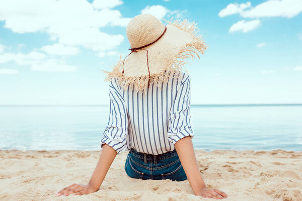 задний вид женщины в соломенной шляпе, отдыхающей на песчаном пляже
 