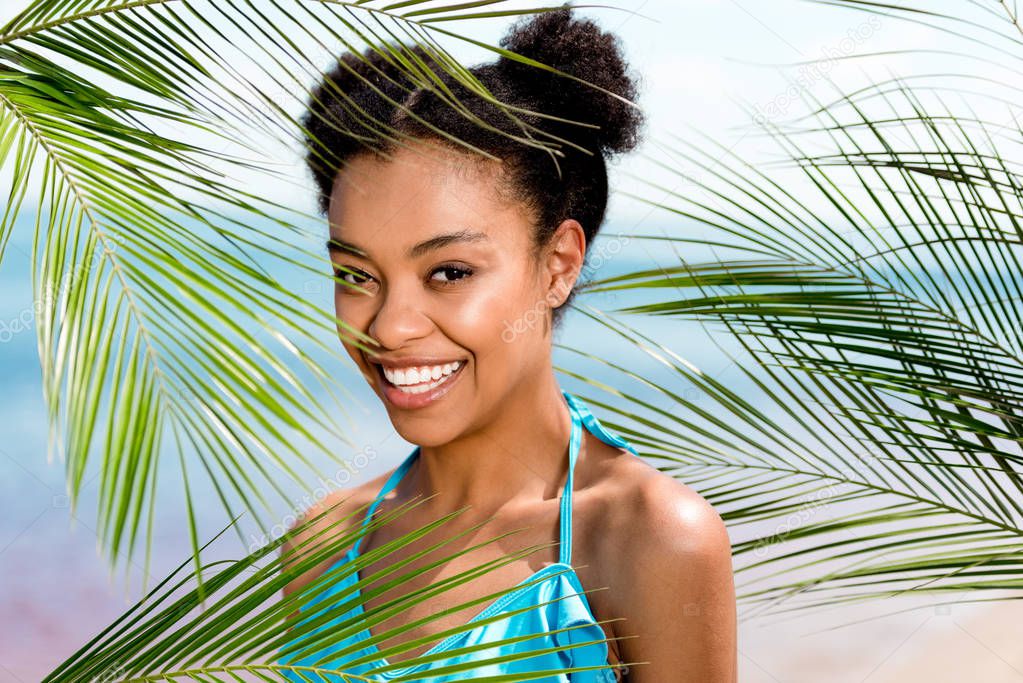 portrait of smiling african american woman near palm leaves in front of sea 