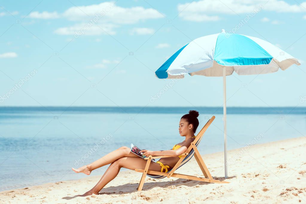 side view of african american woman reading magazine and relaxing on deck chair under beach umbrella in front sea 