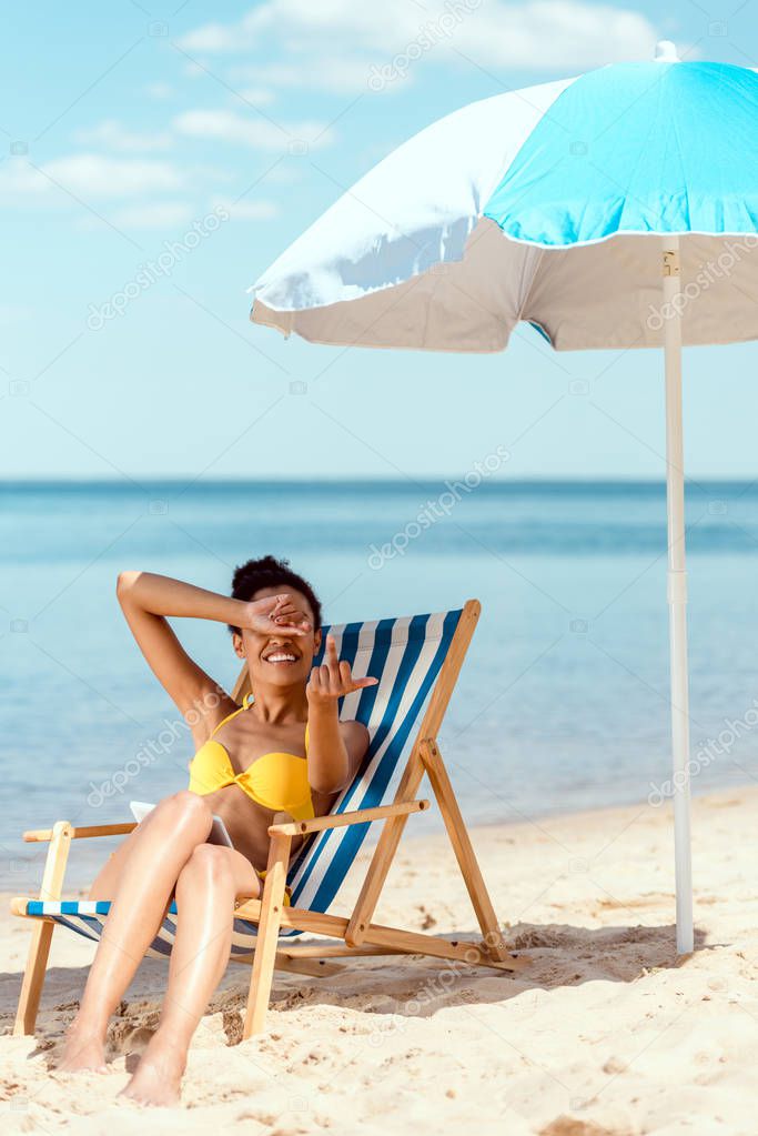 young woman covering eyes by hand and showing middle finger while laying on deck chair under beach umbrella in front of sea 