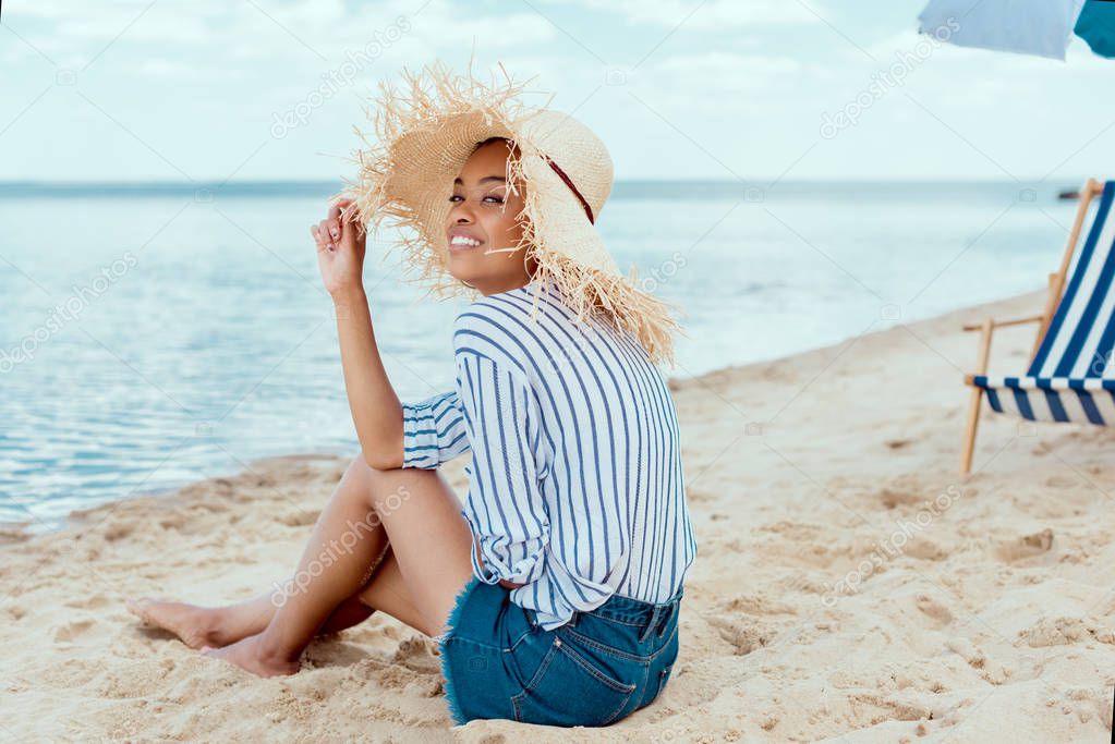 smiling african american woman in straw hat looking at camera while sitting on sandy beach with deck chair and beach umbrella 