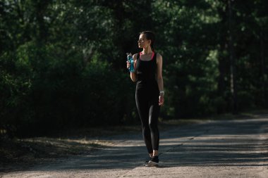 athletic girl holding sports bottle with water and walking in park clipart