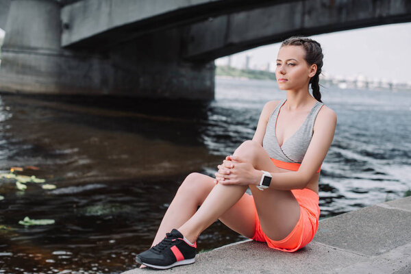attractive sportswoman in sneakers sitting on quay near river