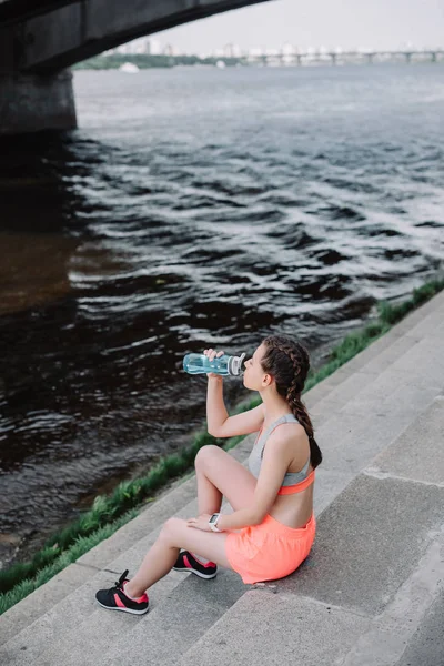 athletic girl drinking water from sports bottle while sitting on quay near river