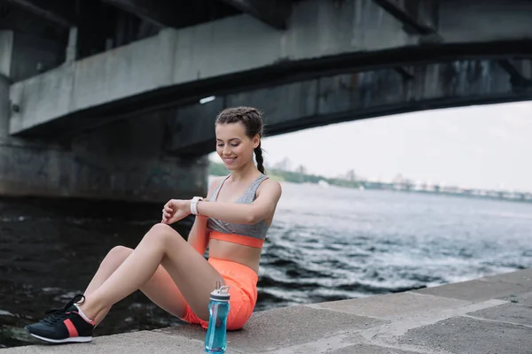 水のボトルと岸壁に座ってスマートな時計を見て笑みを浮かべてスポーツウーマン — ストック写真