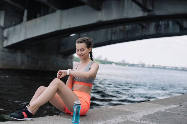 smiling sportswoman looking at smart watch while sitting on quay with bottle of water 