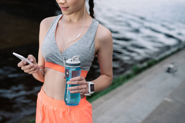 cropped view of young sportswoman holding smartphone with earphones and sports bottle with water
