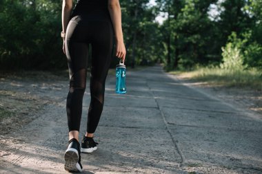 cropped view of sportswoman with sports bottle of water walking on path clipart